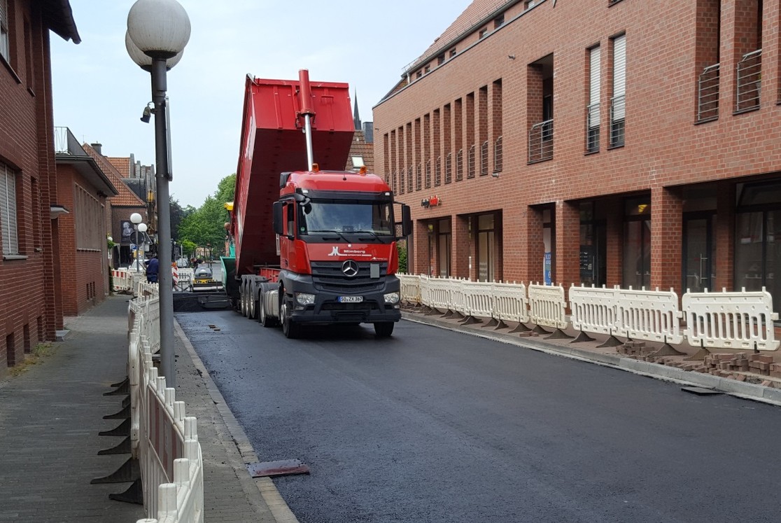 Der Asphaltfertiger schiebt sich langsam über die Straße und verteilt dabei gleichmäßig den Asphalt der Deckschicht über die gesamte Straßenbreite. Die Straßeneinläufe sind abgedeckt. (Bild: Gemeinde Senden)